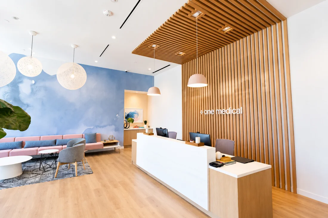 One Medical — Reception under a timber-slat canopy with pink dome pendants, and behind it a blue watercolour wall with woven globe lamps